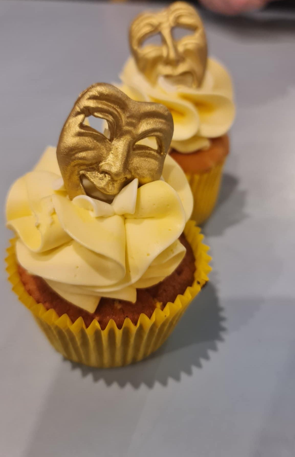 Two vanilla cupcakes with golden theatre mask toppers and soft yellow buttercream swirls in yellow cases